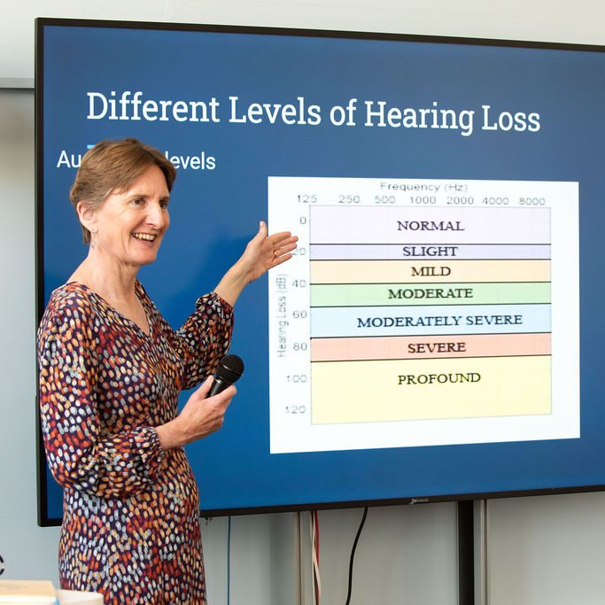 Rachel stood with a microphone, gesturing towards a large screen that shows a diagram on hearing loss.